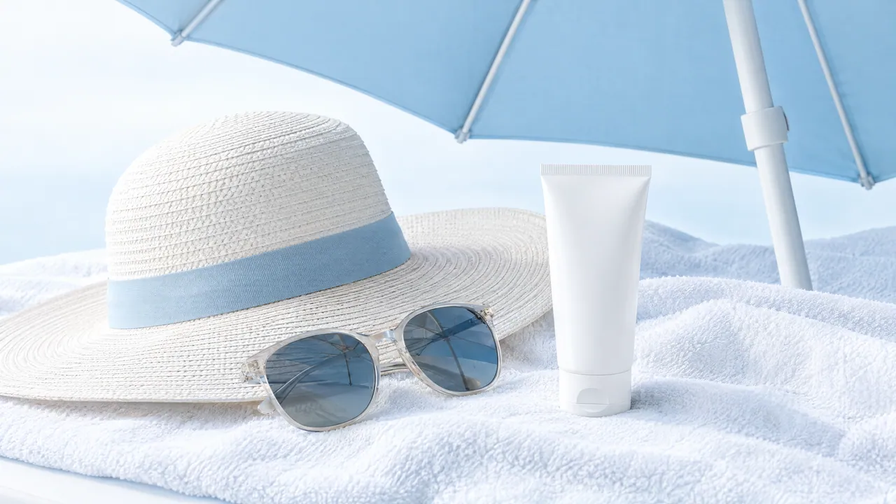 White sun hat, sunglasses and a tube of sunscreen on a white towel under a blue parasol
